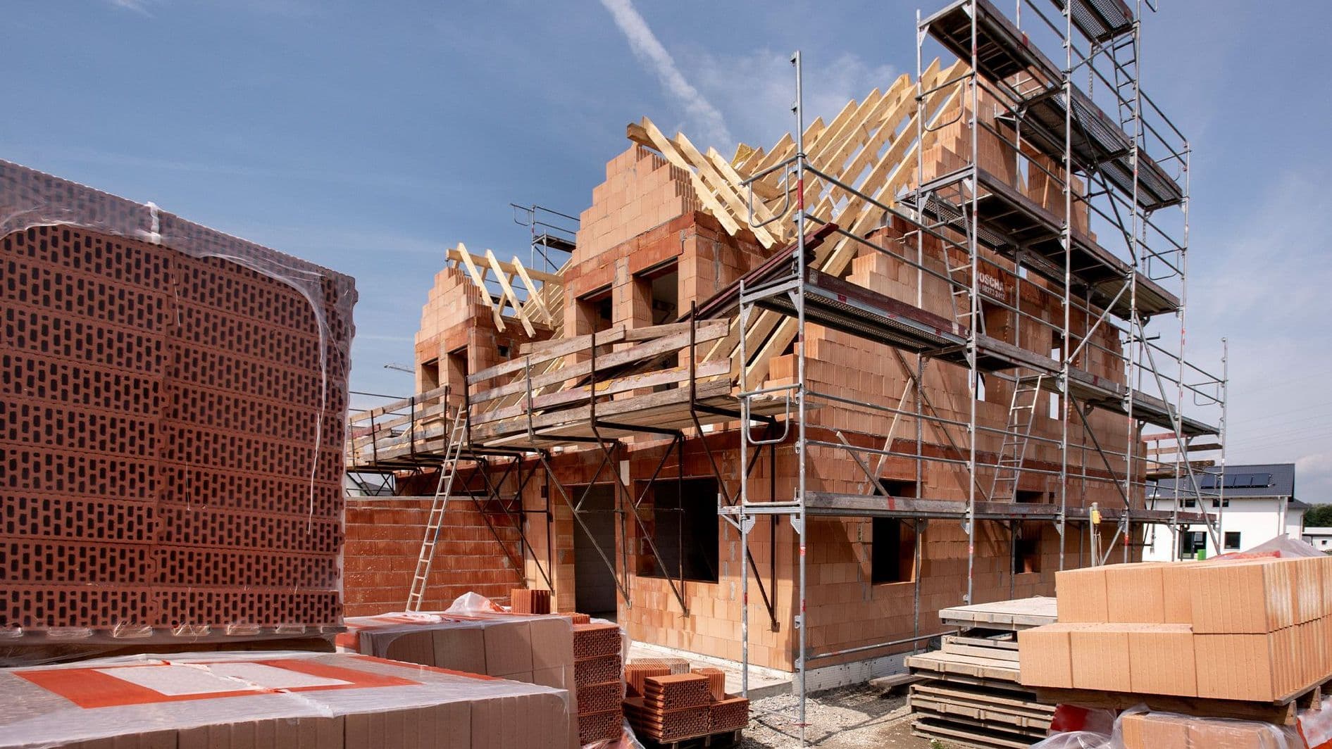 Ein im Bau befindliches Haus aus roten Ziegeln mit Gerüsten an der Fassade. Vor dem Gebäude liegen Stapel von Mauersteinen.