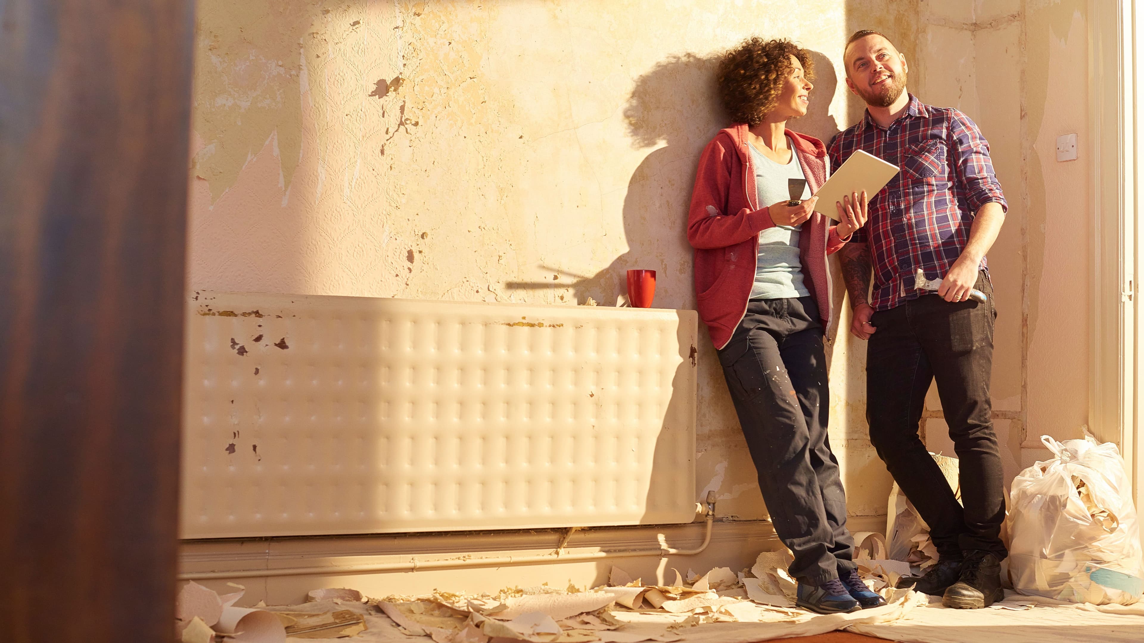 Jung kauft Alt Ein Paar steht in einem Raum mit abblätternder Tapete und Putz. Sie lächeln, halten zufrieden Tablet und Pinsel in der Hand und planen die anstehenden Renovierungsund Modernisierungsarbeiten. Auf dem Boden liegen Tapetenreste, daneben ist ein Müllsack.