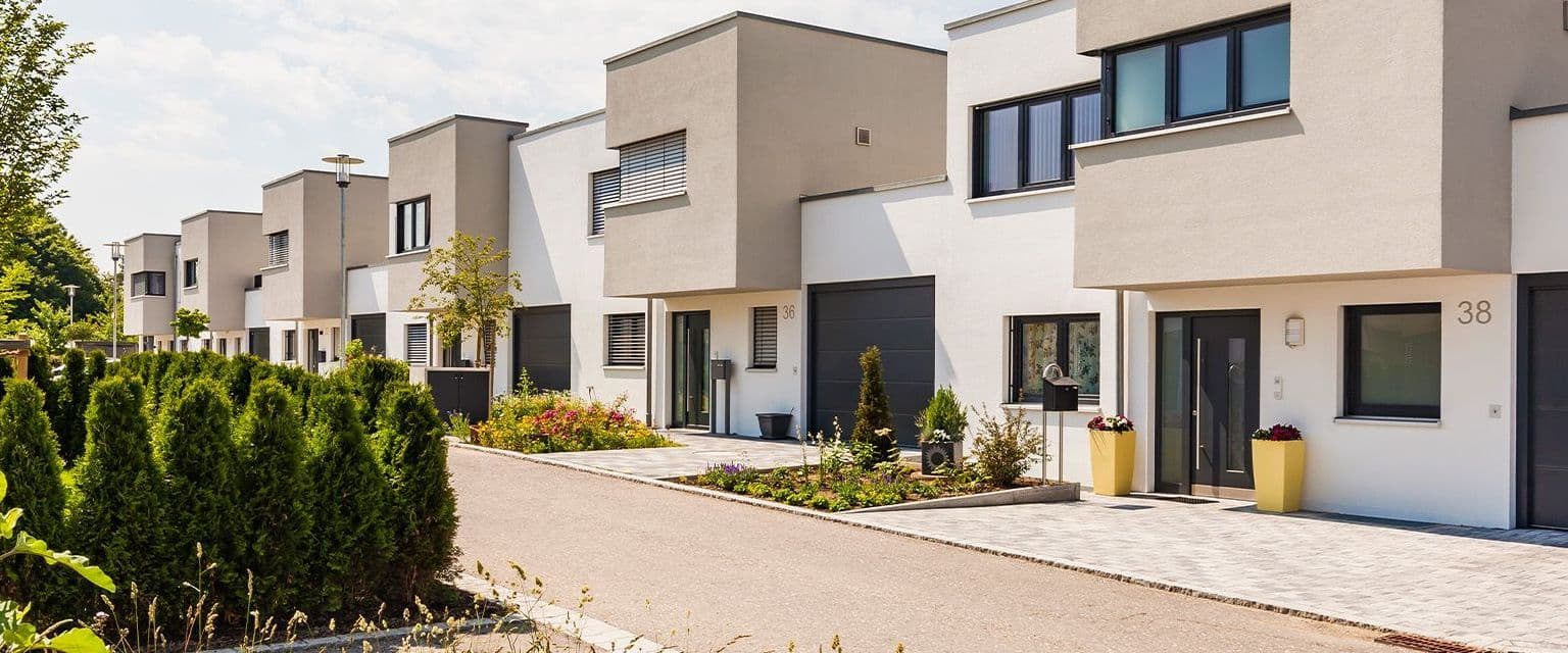 Reihe moderner, zweigeschossiger Stadthäuser mit Vorgärten und Garagen in einer ruhigen Wohnsiedlung. Die Szene zeigt gepflegte Neubauten bei sonnigem Wetter und vermittelt urbanes, familienfreundliches Wohnen.