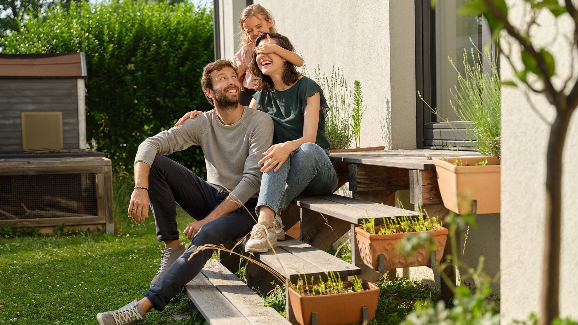 Eine Familie sitzt lachend auf den Eingangsstufen eines Hauses. Ein Mann und eine Frau mit Kind auf dem Arm genießen gemeinsam die Sonne im Garten.