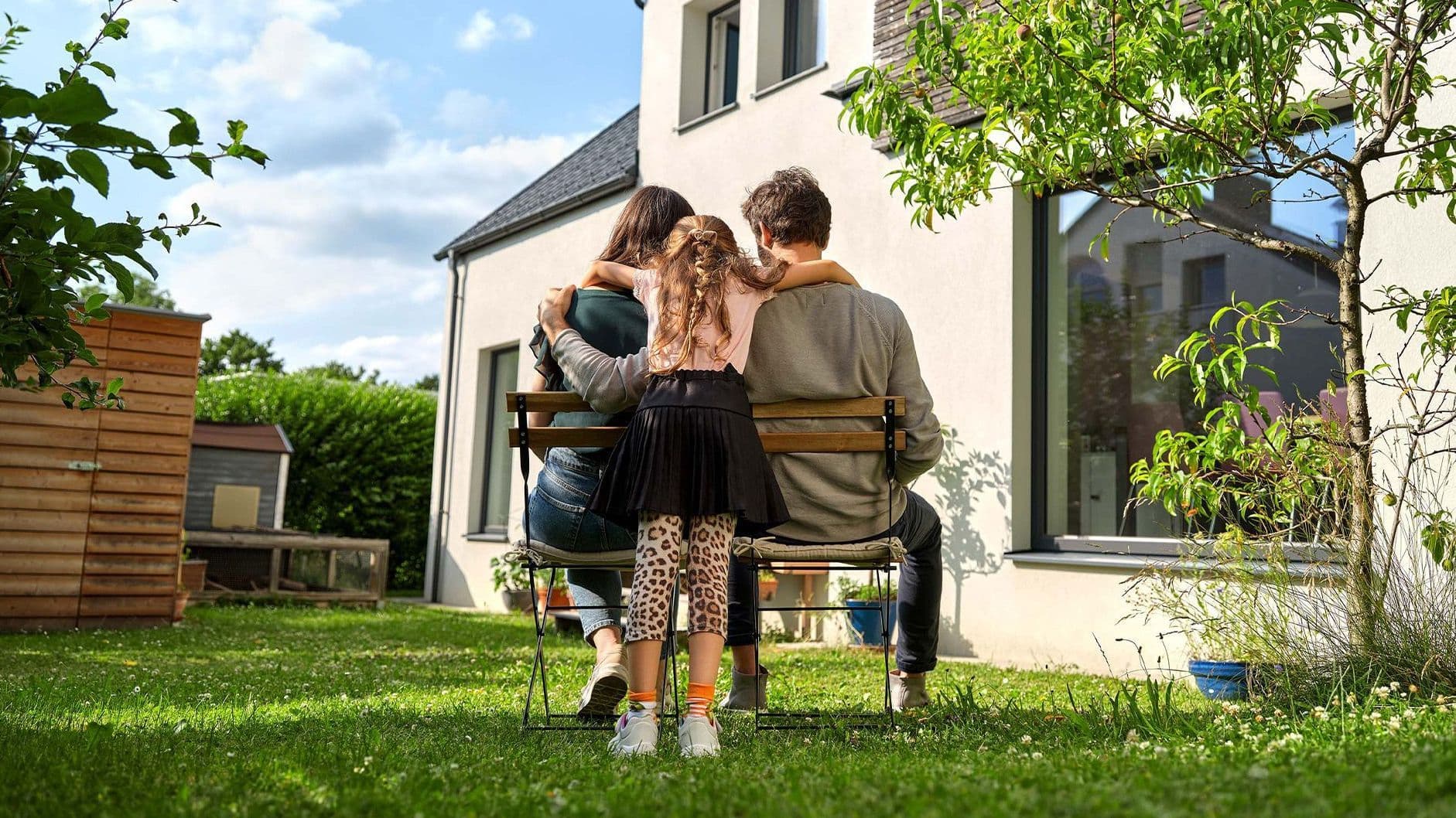 Eine Familie sitzt auf einer Bank im Garten vor einem modernen Haus. Ein Mann, eine Frau und ein Kind umarmen sich von hinten, während sie gemeinsam in den Garten schauen.