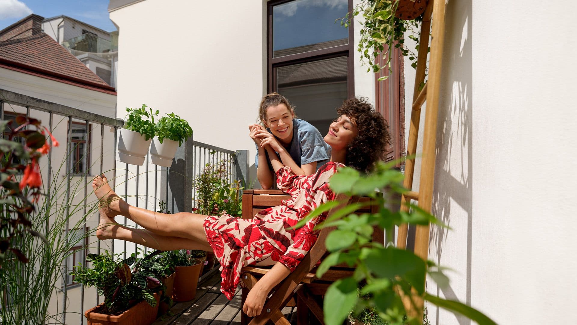 Zwei Personen sitzen entspannt auf einem Balkon mit vielen Pflanzen. Eine Person trägt ein rotes Kleid und lehnt sich zurück, während die andere sich lächelnd auf das Geländer stützt.