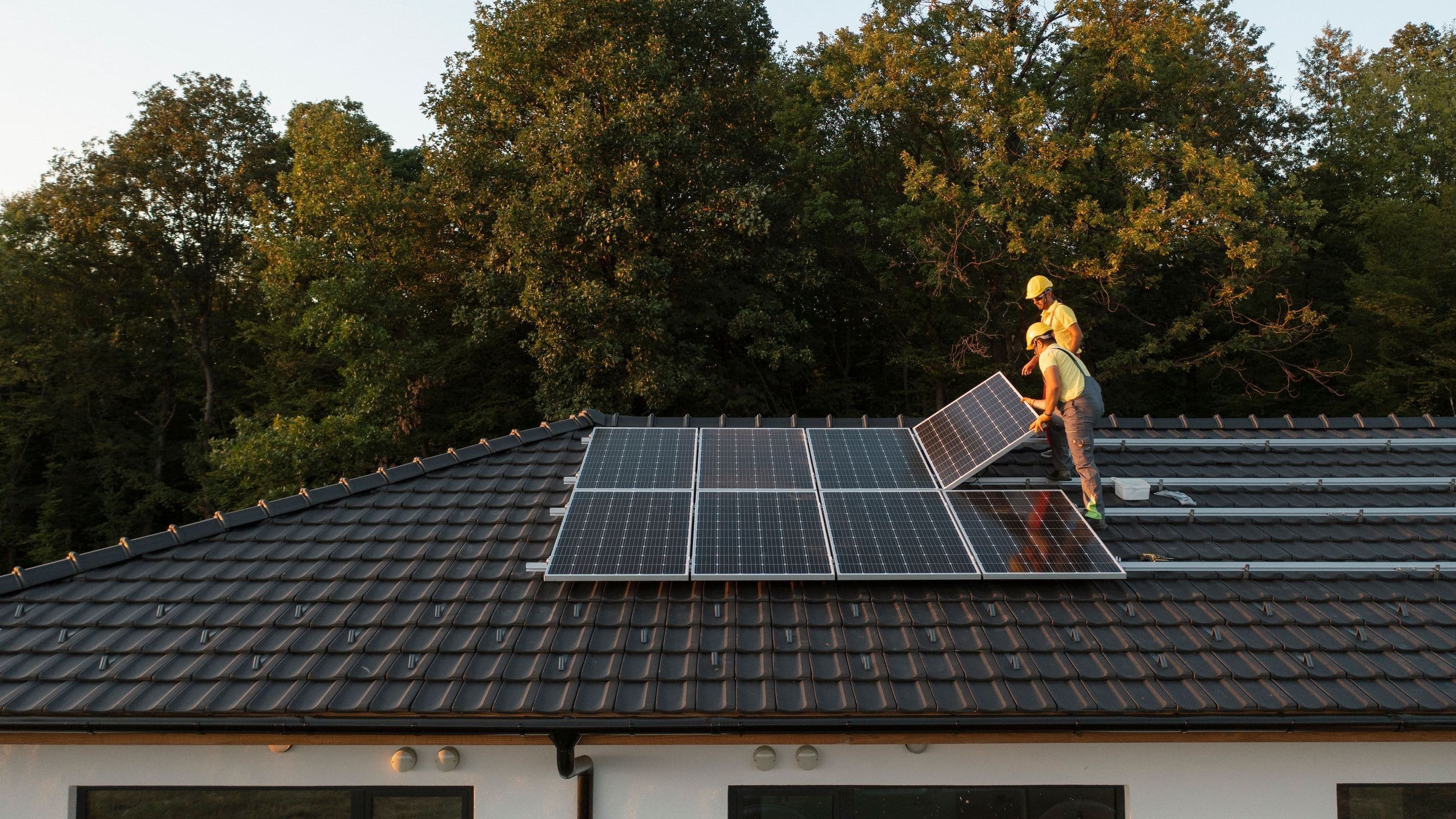 Zwei Handwerker installieren Solarmodule auf dem Dach eines Einfamilienhauses. Sie tragen Schutzhelme und arbeiten im Abendlicht, während hinter dem Haus Bäume im Sonnenuntergang leuchten.