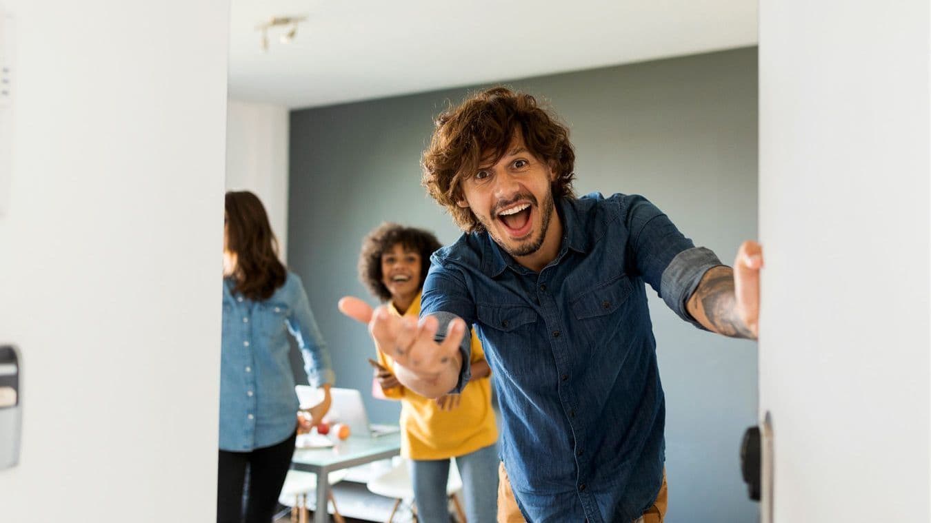 Lächelnder Mann mit Bart und lockigen Haaren öffnet fröhlich die Tür, während zwei Personen im Hintergrund ebenfalls lächeln.