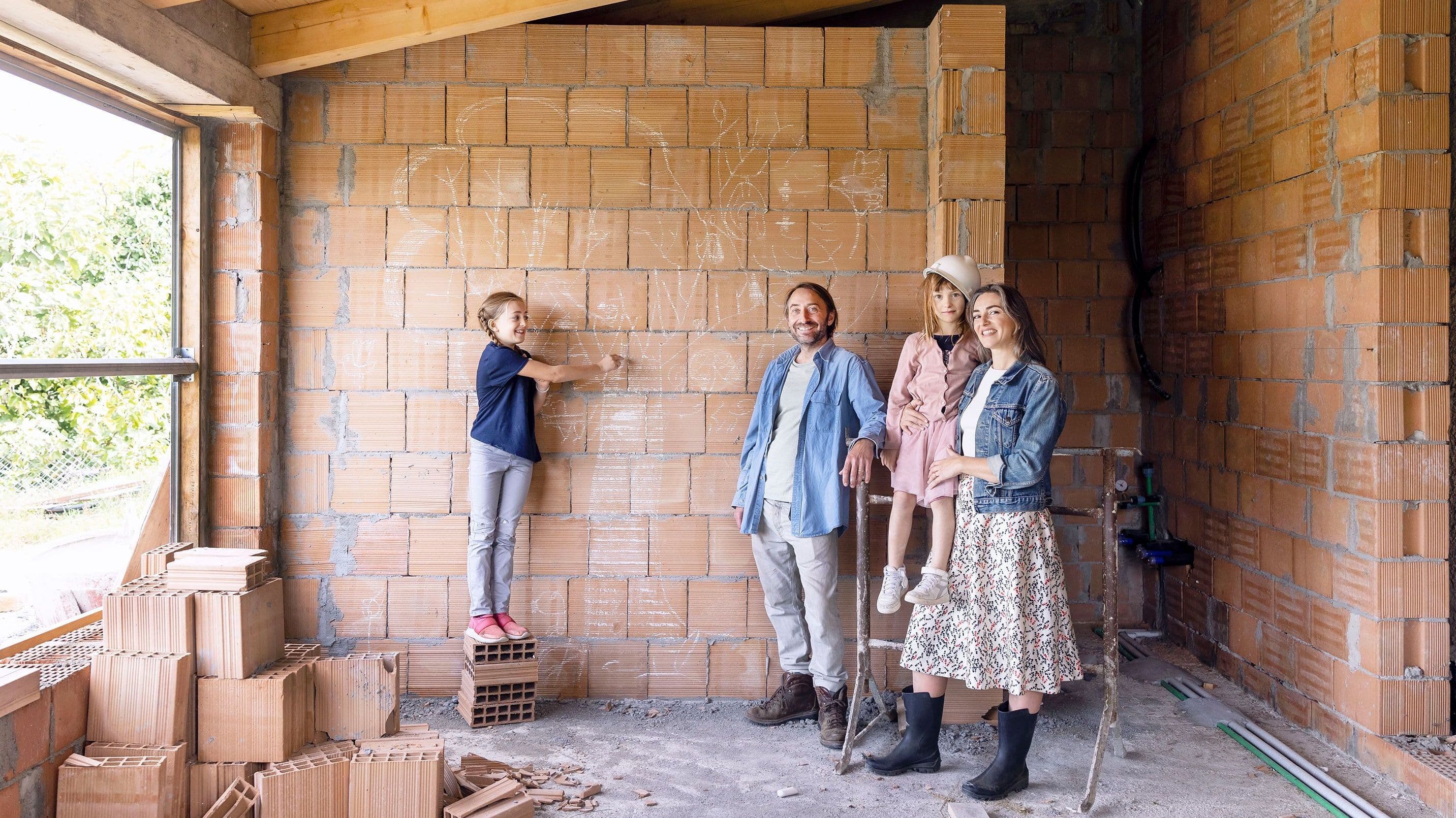 Eine vierköpfige Familie steht in dem Rohbau ihres neuen Hauses vor einer unverputzten Mauersteinwand und schaut in die Kamera.