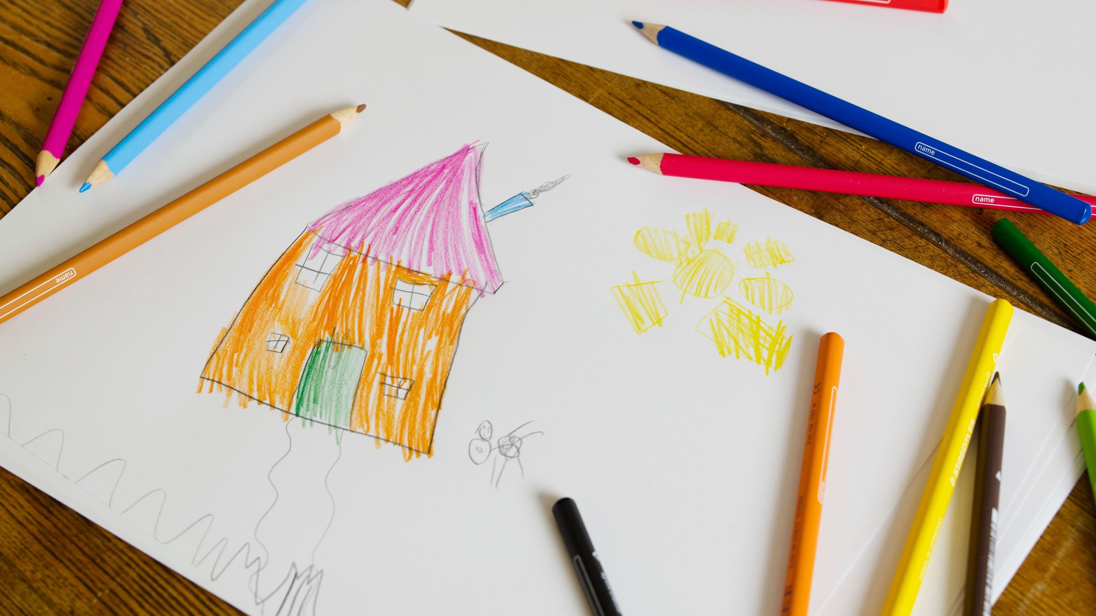 Ein Kinderzeichnung liegt mit Buntstiften auf einem Holztisch. Die Zeichnung zeigt ein Haus und eine Sonne.