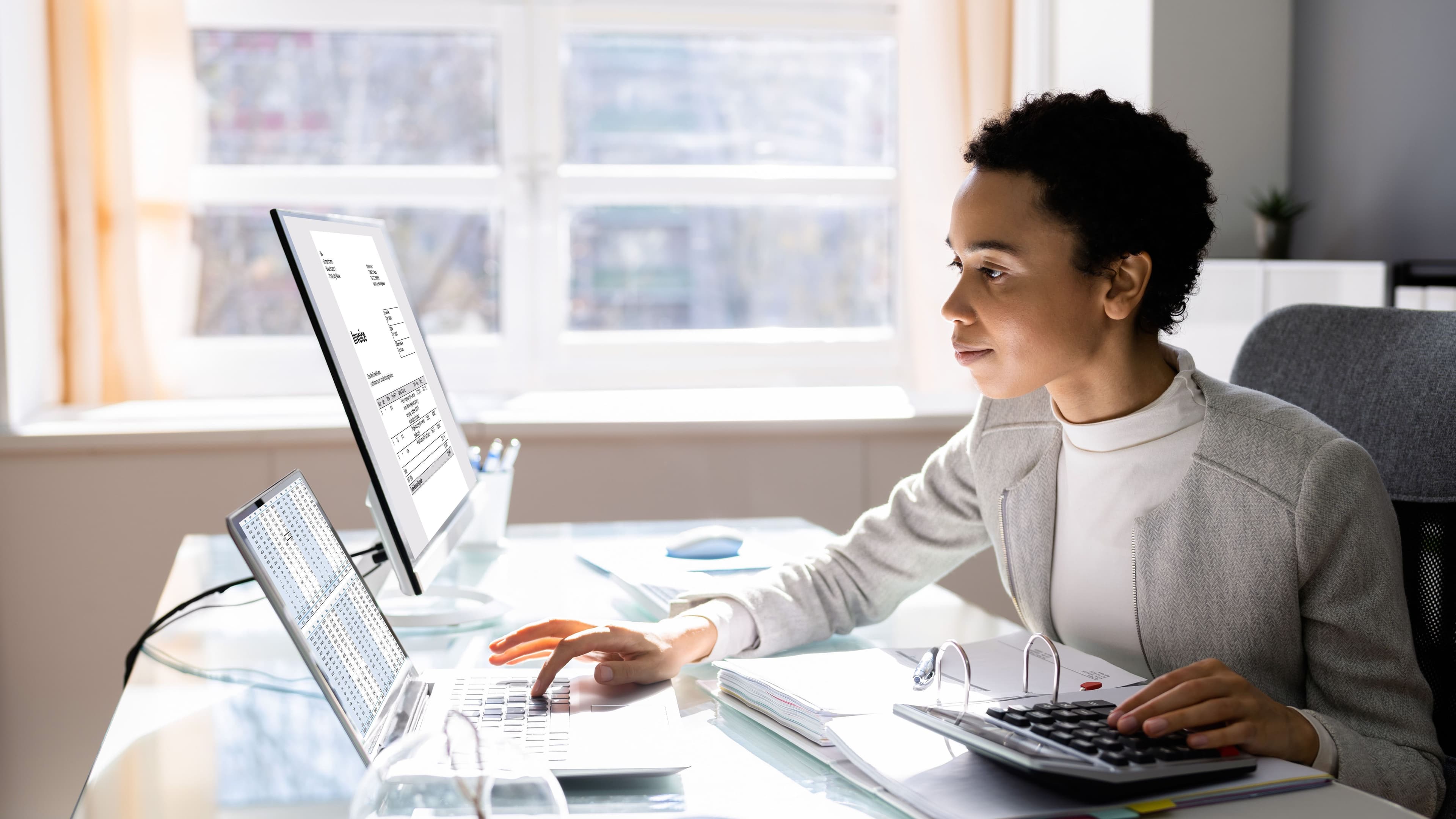 Eine Frau sitzt mit einem Rechner vor einem Laptop und einem Bildschirm. Mit der linken Hand bedient sie den Rechner, mit der rechten Hand tippt sie am Laptop.