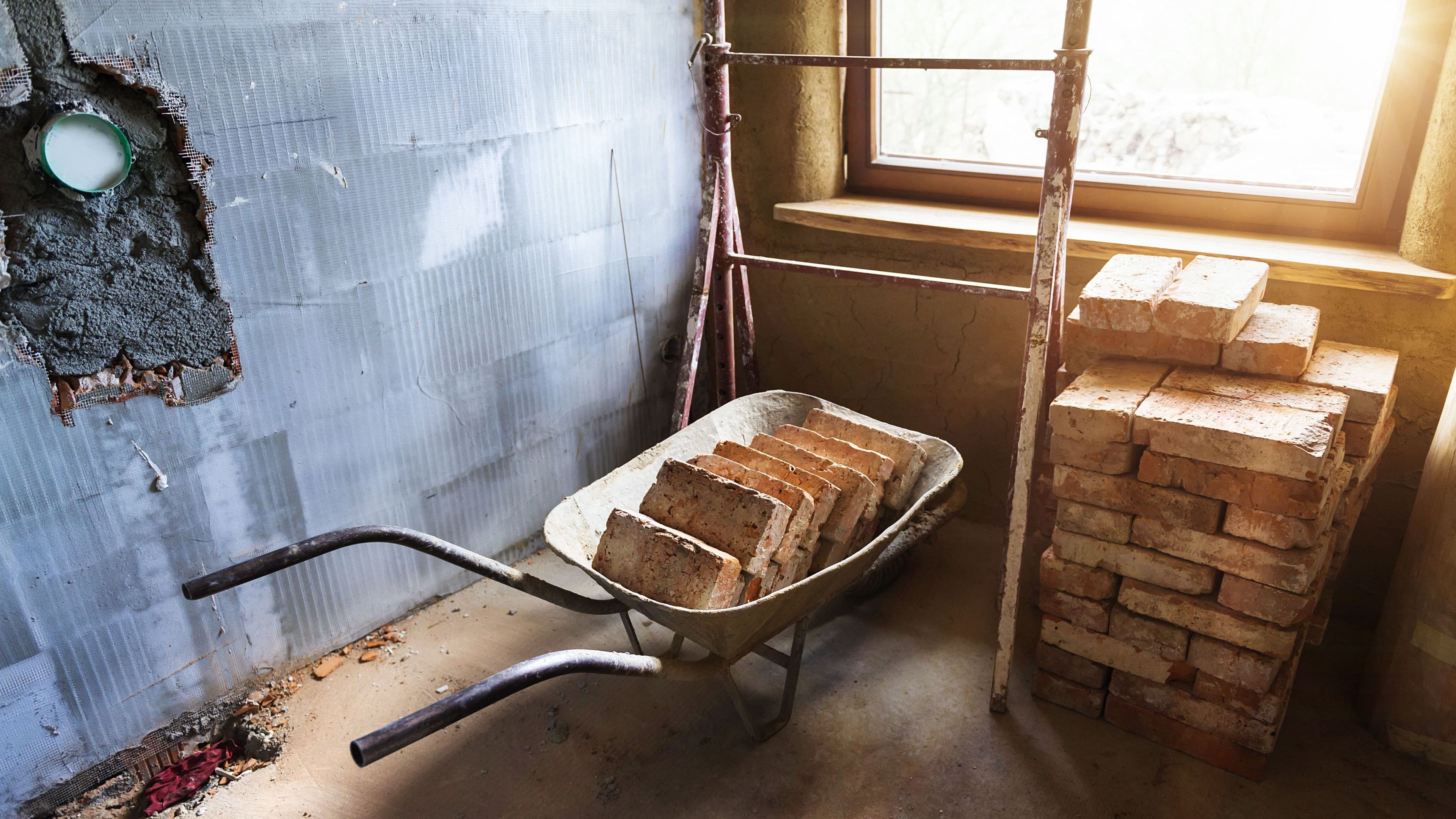 Eine Schubkarre mit Ziegelsteinen steht auf einer Baustelle vor einem Fenster. Daneben liegen weitere Ziegelsteine aufgestapelt.